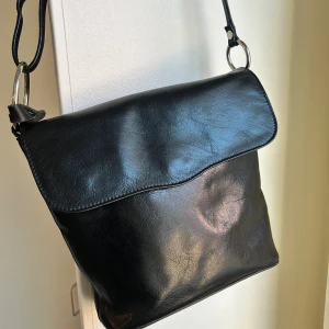 Skinnväska Wera  - Axelremsväska i skinn från Wera med justerbar axelrem.  Har ett mittfack med dragkedja ett innerfack som stängs med dragkedja.  • Axelväska i skinn • Stilren design • Justerbar axelrem • Stängning med magnetknapp  Mått: Längd 27cm Höjd 26cm Djup 7cm