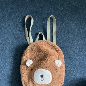 Barnväska  - Hej! Säljer en brun/ begie väska för barn med björn utseende, kommer från H&M o är i mycket fint skick, ett stort fack med dragkedja o ett litet utan dragkedja🤍