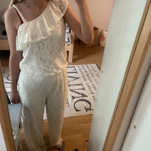 Vit byxdress med volangdetalj - Säljer en superfin vit spets byxdress med en snygg volangdetalj över axeln. Byxdressen är ärmlös. Perfekt på sommaren den är använd 1 gång så är även i bra skick. Storlek 158/164 men passar xs-s