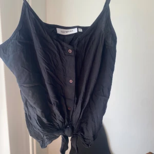 Svart knytblus från Size Needle - Säljer en snygg svart blus från Size Needle i storlek M. Den har knappar framtill och en cool knytning längst ner. Perfekt för en avslappnad sommarlook!