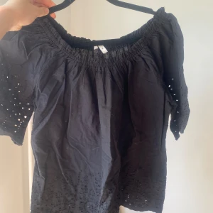 Svart offshoulder topp - Säljer en snygg svart offshoulder topp med broderade detaljer. Toppen har korta ärmar och en luftig passform, perfekt för sommardagar. Den är i bomull och känns väldigt bekväm att ha på sig.