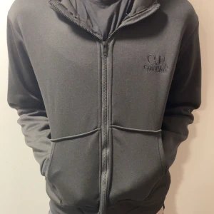 Grå hoodie från C.P. Company - Säljer en stilren grå hoodie från C.P. Company med dragkedja och coola glasögondetaljer på huvan. Perfekt för en casual look. Den har långa ärmar och en normal passform. PRISET KAN DISKUTERAS‼️