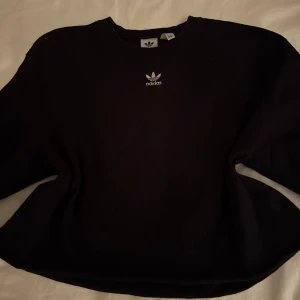 Svart croppad tröja från Adidas - Säljer en svart croppad tröja från Adidas med deras ikoniska logga på bröstet. Tröjan har en rund halsringning och är perfekt för träning. Passar bra till träningsbyxor.