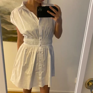 Vit kortklänning med knappar - Säljer en söt vit kortklänning med knappar framtill och en smickrande resår i midjan. Perfekt för varma sommardagar. Klänningen har en krage och korta ärmar som ger en avslappnad look. Passar perfekt för en dag på stan eller en picknick i parken.