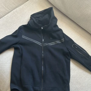 Svart hoodie från Nike - Säljer en svart hoodie från Nike med dragkedja och huva. Jackan har en reflekterande rand över bröstet och en praktisk ficka med dragkedja på ärmen. Perfekt för en sportig look eller avslappnade dagar.