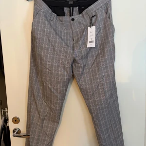 Grå rutiga kostymbyxor - Snygga grå kostymbyxor med ett klassiskt rutmönster. Byxorna har en rak passform och är perfekta för en stilren look. De har knappar och dragkedja framtill. Storlek 34 