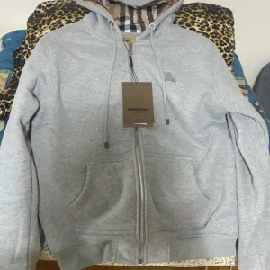 Grå huvtröja från Burberry - Säljer en stilren grå huvtröja från Burberry med dragkedja och broderad logga på bröstet. Insidan av huvan har det klassiska Burberry-mönstret. Perfekt för en avslappnad look.