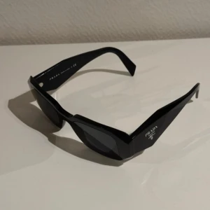Svarta solglasögon från Prada - Snygga svarta solglasögon från Prada med en modern och stilren design. Tillverkade i Italien, dessa glasögon har en bred båge och mörka linser som ger ett elegant intryck. Perfekta för att ge din outfit en lyxig touch.