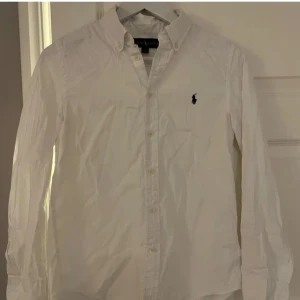 Vit skjorta från Ralph Lauren - Säljer en klassisk vit skjorta från Ralph Lauren med broderad logga på bröstet. Skjortan har långa ärmar och knappar framtill. Perfekt för en stilren look.sitter bra runt 160 till 170 beroende på vikt också. Har du någon fråga är de bara att fråga på