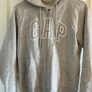 Grå hoodie från GAP - Säljer en klassisk grå hoodie från GAP med dragkedja och justerbar huva. Tröjan har ett stort GAP-tryck på framsidan och är tillverkad i mjukt material för extra komfort. Perfekt för en avslappnad stil.