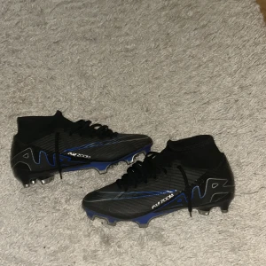 Svarta och blå fotbollsskor från Nike - Snygga svarta fotbollsskor från Nike med blå och silver detaljer. Skorna har snörning och en hög ankelkrage för extra stöd. Perfekta för fotbollsplanen med sin moderna design och funktionalitet.