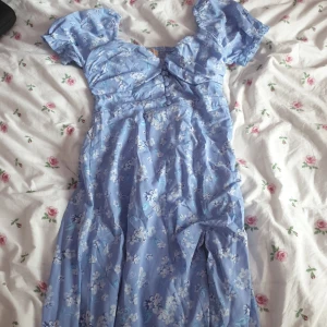 Blommig blå klänning - Charmig blå klänning med blommigt mönster och korta ärmar. Klänningen har en smickrande passform med knappar framtill och en lätt rynkad design. Perfekt för somriga dagar.