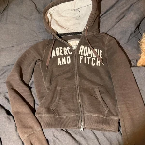 Brun hoodie från Abercrombie & Fitch - Säljer en brun hoodie från Abercrombie & Fitch med dragkedja och huva. Tröjan har långa ärmar och en stor logga på framsidan. Dragkedjan är dock trasig, så går ej att stänga tjocktröjan. Den ör otroligt snygg och sitter som en smäck! På tröjan står det storlek L men skulle säga att den sitter som en tajt M eller S.