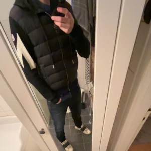 Svart dunjacka från Moncler - Snygg svart cardigan från Moncler med dragkedja och huva. Cardiganen har ett distinkt vitt mönster på ärmen och Moncler-logotypen på axeln. Perfekt nu för våren. Det är en sällsynt och ovanlig cardigan. Dragkedjan kan krångla ibland men fungerar perfekt om man lär sig. Storlek M, pris kan diskuteras. Hör av er om det minsta mvh Axel. 