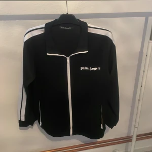 Svart tröja Palm Angels - Snygg svart track jacket från Palm Angels med vita ränder längs ärmarna och dragkedja framtill. Jackan har en klassisk look med broderad logga på bröstet. Perfekt för en stilren och sportig outfit.