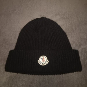 Svart ribbad mössa från Moncler - Snygg svart ribbad mössa från Moncler med deras ikoniska logotyp framtill. Perfekt för att hålla värmen under kyliga dagar. Stilren design som passar till det mesta.