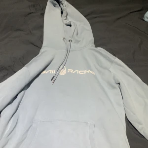 Ljusblå hoodie från Sail Racing - Säljer en ljusblå hoodie från Sail Racing med tryck på bröstet. Den har en klassisk känguruficka och justerbar huva med dragsko. Perfekt för en avslappnad stil.
