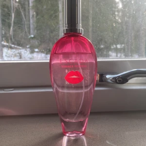 Escada Summer Festival Parfym - Säljer en Escada Summer Festival parfym i en elegant rosa flaska med silverfärgad kork. Flaskan har en stilren design med ett tryck av röda läppar på framsidan. Perfekt för att ge en fräsch och somrig doft.