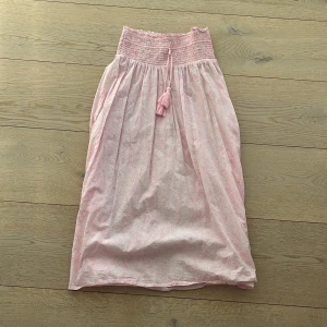 Rosa kjol från Gina Tricot - Söt rosa kjol från Gina Tricot med smockdetaljer i midjan och dekorativ tofs. Kjolen har ett diskret mönster och är perfekt för varma dagar. Den är lätt och luftig, vilket gör den bekväm att bära. Passar bra till en avslappnad stil.