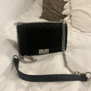 Svart axelväska med kedja - Snygg svart axelväska med quiltat mönster och silverfärgad kedja. Väskan har en praktisk dragkedja inuti och ett stilrent spänne framtill. Perfekt för att ge en elegant touch till din outfit.