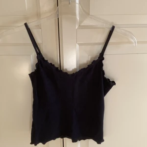 Svart ribbad linnetopp från Hollister  - Säljer en söt svart ribbad linnetopp från Hollister. Toppen har smala axelband och en vågig kantdetalj vid halsringningen och nederkanten. I fint skick. Kort modell. Storlek L, men passar M.