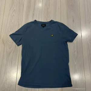 Blå t-shirt från Lyle & Scott - Säljer en snygg blå t-shirt från Lyle & Scott Junior. Den har en klassisk design med korta ärmar och ett broderat gult emblem på bröstet. Perfekt för en stilren och avslappnad look. Bra pris 👀