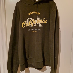 Grön hoodie med tryck - Säljer en grön hoodie med tryck i vitt och gult. Texten 'California' och 'Denim Goods' ger en vintagekänsla. Hoodien har en klassisk passform med långa ärmar och en justerbar huva med snören.
