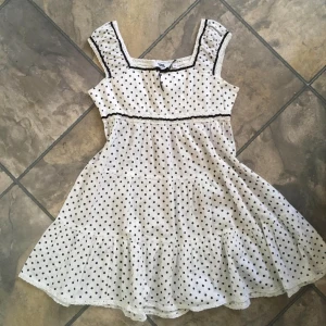 Jätte fin Vit prickig klänning från Heartquake Lindex - Charmig vit klänning med svarta prickar från Heartquake. Klänningen har en söt rosett framtill och svarta spetsdetaljer vid ärmar och midja. Perfekt för en somrig look med sin luftiga design och ärmlösa stil. 🖤Knappt använd så i Super fint skick 🌟