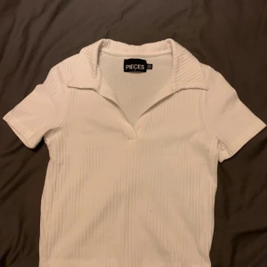 Vit ribbad polotopp från Pieces - Säljer en stilren vit ribbad polotopp från Pieces. Toppen har korta ärmar och en klassisk krage. Perfekt för en enkel och snygg look.