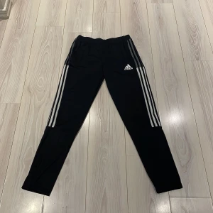Svarta träningsbyxor från Adidas - Säljer ett par svarta träningsbyxor från Adidas med de klassiska tre ränderna längs sidorna. Byxorna har en elastisk midja och är tillverkade i ett lätt och bekvämt material. Nypris 350kr Perfekta för sport och träning, byxorna är i nyskick. Billigt och bra pris!!!👀