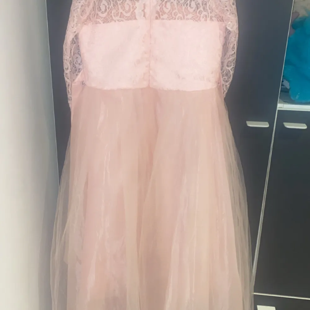 Elegant ljusrosa klänning med spetsdetaljer och trekvartsärmar. Klänningen har en vacker tyllkjol och en söt rosett i midjan. Perfekt för speciella tillfällen.. Mekot.
