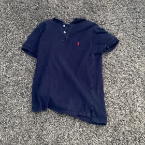 Mörkblå pikétröja från Ralph Lauren - Säljer en klassisk mörkblå pikétröja från Ralph Lauren med röd broderad logga på bröstet. Tröjan har korta ärmar och knappar vid halsen. Perfekt för en stilren look.