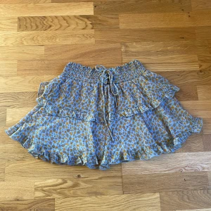 Blommig kortkjol med volang - Charmig blommig kortkjol från STR i blått och orange. Kjolen har en volangdetalj och ett knytband i midjan för justerbar passform. Perfekt för en somrig look!