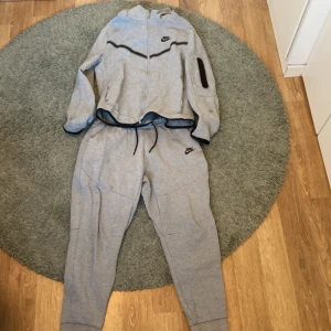 Grå träningsset från Nike - Säljer en grå Nike tech dress  från Nike med dragkedja och svarta detaljer. Setet består av en hoodie med lång ärm och matchande byxor med dragsko i midjan. Perfekt för träning eller en chill dag.skicket är på 8/10 för att ett litet hål på armen men det syns inte. Kan gå ner till Max 600kr+frakt