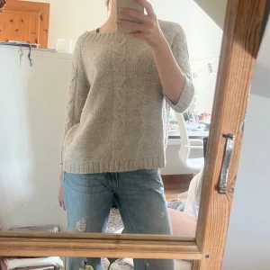 Beige stickad tröja - Säljer en mysig beige stickad tröja med kabelstickat mönster framtill. Tröjan har trekvartsärmar och en avslappnad passform. Perfekt för skolan men även finare tillfällen då man kan ha många synliga smycken till. Storleken är M men passar även på S och XS