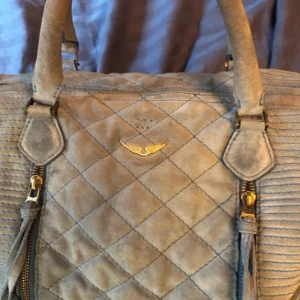 Beige quiltad väska från Zadig & Voltaire - Snygg beige väska från Zadig & Voltaire i quiltat mönster. Väskan har detaljer som dragkedja och fransar, vilket ger den en unik look. Perfekt för dig som vill ha en stilren och trendig accessoar.