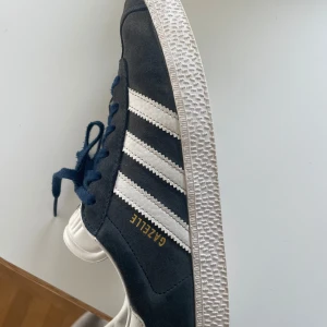 Adidas Gazelle sneakers i märkblå och vitt - Snygga Adidas Gazelle sneakers i mörkblå mocka med vita detaljer och blå snörning. De har den klassiska tre-randiga designen på sidorna och en vit sula. Perfekta för en stilren och sportig look.