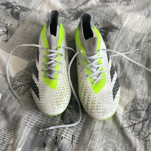  Adidas Predator AG och FG Elite  - Snygga fotbollsskor i vitt med neongula och svarta detaljer. Skorna har snörning och en modern design som passar perfekt för fotbollsplanen.