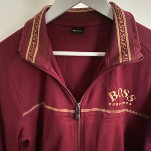 Vinröd kofta från Hugo Boss - Säljer en snygg vinröd jacka från Hugo Boss med dragkedja och broderad logga på bröstet. Jackan har långa ärmar och ribbad krage med märkets namn längs kanten. Perfekt för en stilren look.