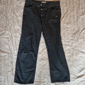 Svarta jeansbyxor från H&M - Snygga svarta jeansbyxor från H&M med en avslappnad passform. Byxorna har en knappgylf och slitna detaljer på ena benet för en trendig look. Perfekta för en casual stil. Ankellånga