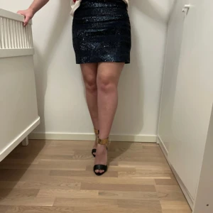 Svart/blå glittrig kortkjol - Snygg svart glittrig kortkjol perfekt för en utekväll. Kjolen har en tight passform som framhäver figuren och ger en elegant look. Den är enkel att matcha med olika toppar och accessoarer för en komplett outfit.