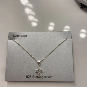 Halsband i 925 Sterling Silver med Korshänge - Elegant halsband i 925 sterling silver med ett korshänge dekorerat med glittrande stenar. Perfekt för att ge en stilren touch till din look. Kedjan är tunn och smidig, vilket gör den bekväm att bära.