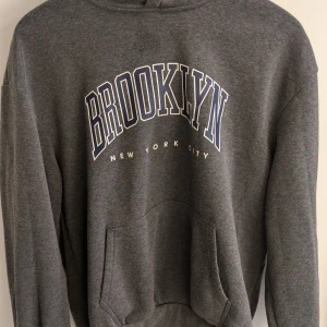 17883 - Säljer en grå hoodie med texten 'Brooklyn New York City' tryckt på framsidan. Tröjan har en klassisk känguruficka och en bekväm huva. Perfekt för en avslappnad stil. Paket storlek 12 gram