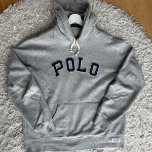 Grå hoodie från Polo Ralph Lauren - Säljer en klassisk grå hoodie från Polo Ralph Lauren med stor POLO-text på bröstet. Den har en praktisk magficka och justerbar huva med snören. Den är i storlek large och passar large. Har ett litet hål på magfickan som syns på bilden. Hör gärna av dig vid frågor. Priset är inte hugget i sten och byten i strl L kan vara intressanta.