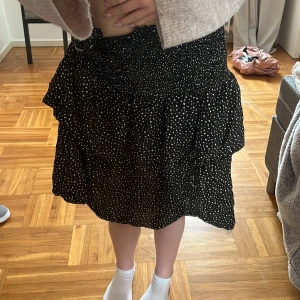 Svart prickig kjol med volang - Säljer en svart kjol med vita prickar och volangdetaljer. Kjolen har en kort längd och en bekväm passform. Perfekt för att ge en lekfull touch till din outfit! Förstora åt mig därför knutit dom