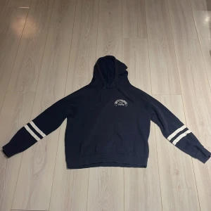 Mörkblå hoodie från Gina Tricot - Säljer en mörkblå hoodie från Gina Tricot med texten 'She is a champion since 1982' på bröstet. Tröjan har långa ärmar med vita ränder och en klassisk huva med dragsko. Perfekt för en avslappnad stil.