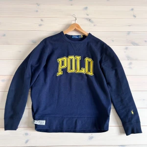 Mörkblå sweatshirt från Polo Ralph Lauren - Säljer en snygg mörkblå sweatshirt från Polo Ralph Lauren med stort gult 'POLO' tryck på framsidan. Tröjan har långa ärmar och en liten logga på ärmen. Perfekt för en avslappnad stil. Skick 9/10