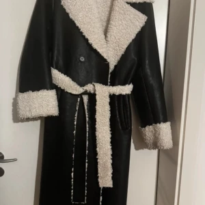 Svart lång kappa med pälsdetaljer - Snygg svart lång kappa med vita pälsdetaljer och bälte i midjan. Perfekt för kyliga dagar med sin stilrena design och varma material. Kappan har knappar framtill och långa ärmar med päls vid ärmsluten. En knapp som fattas men påverkar ingen passar s/m