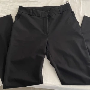 Svarta kostymbyxor - Snygga svarta kostymbyxor med en klassisk design. De har en rak passform och är perfekta för en stilren look. Byxorna har hällor för bälte och en diskret dragkedja framtill.