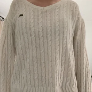 Vit stickad tröja från Lacoste - Säljer en stilren vit stickad tröja från Lacoste med det klassiska krokodilmärket på bröstet. Tröjan har ett flätat mönster och långa ärmar, perfekt för kyligare dagar. Passar både till jeans och kjol för en avslappnad look.